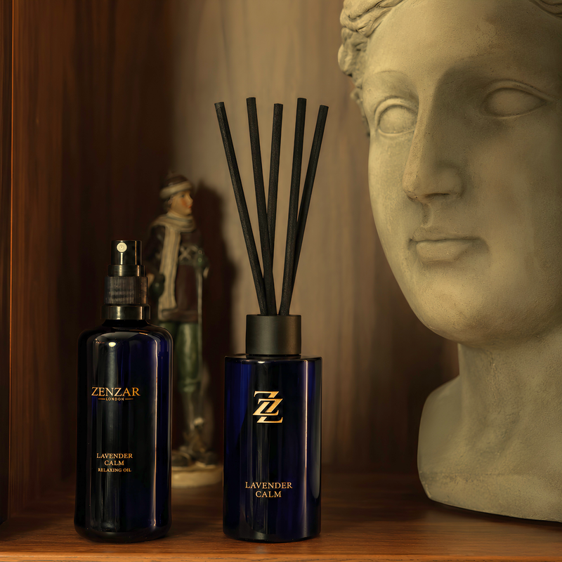 Two ZENZAR Lavender Calm fragrance products displayed on a wooden shelf: a dark blue spray bottle on the left and a matching reed diffuser with black reeds on the right, styled beside a classical bust and small figurine.